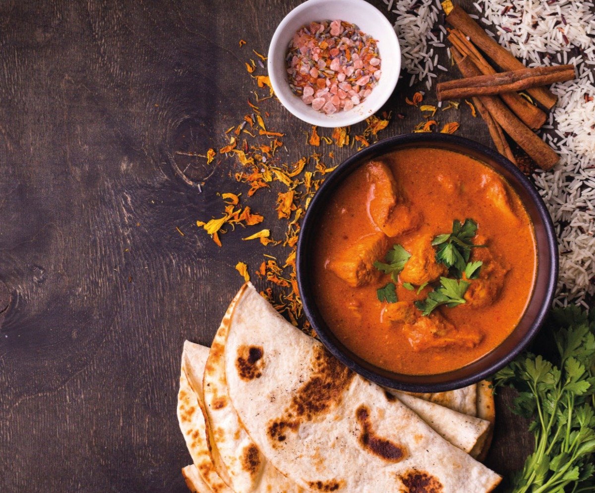 Bowl of Chicken curry with naan on the side, table garnished with cinnamon sticks, pink rock salt, coriander and rice